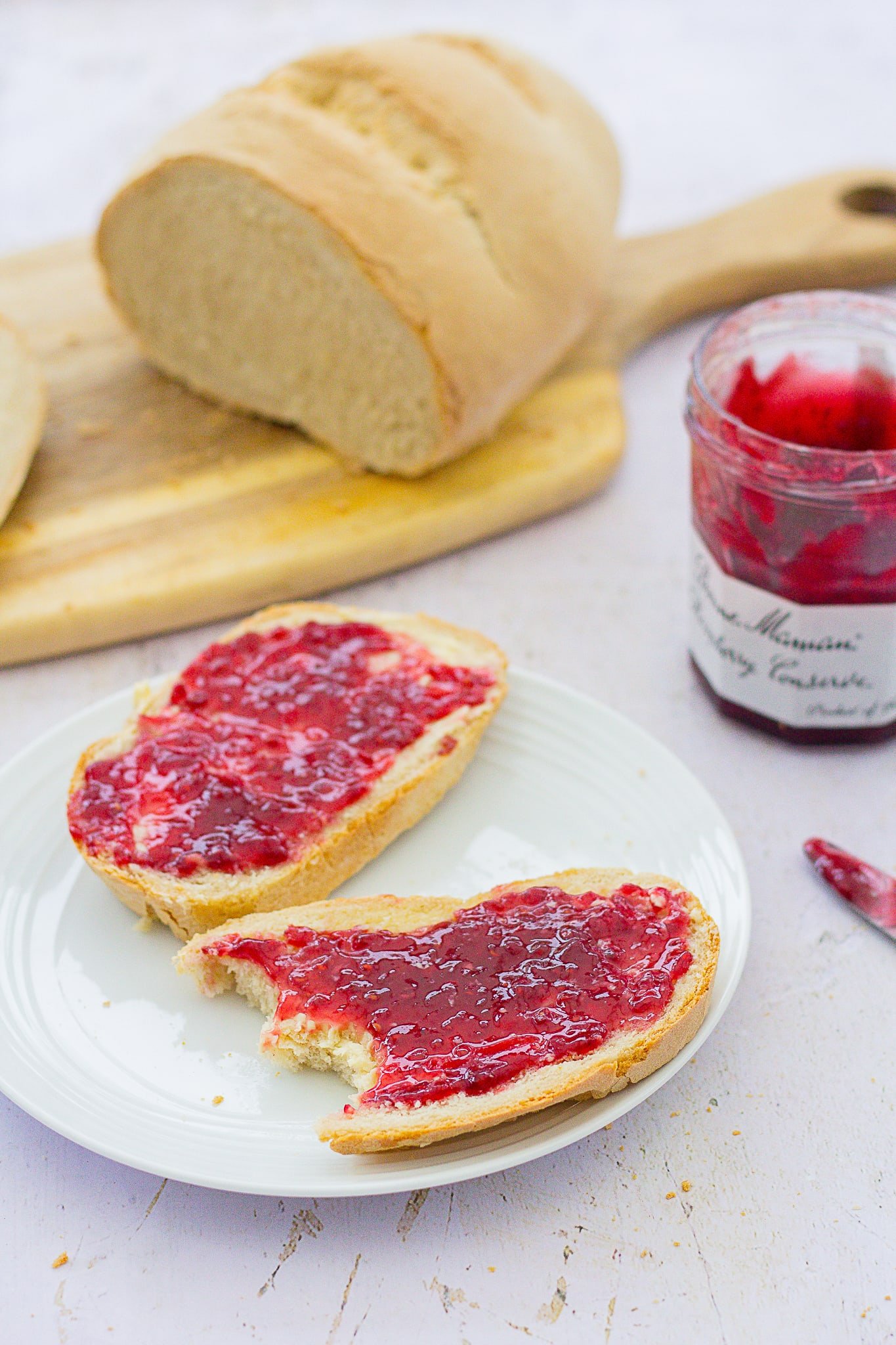 Super Simple White Bread on a board, several slices have been cut off, two slices are on a plate in front covered in raspberry jam, a pot of raspberry jam is next to the plate.