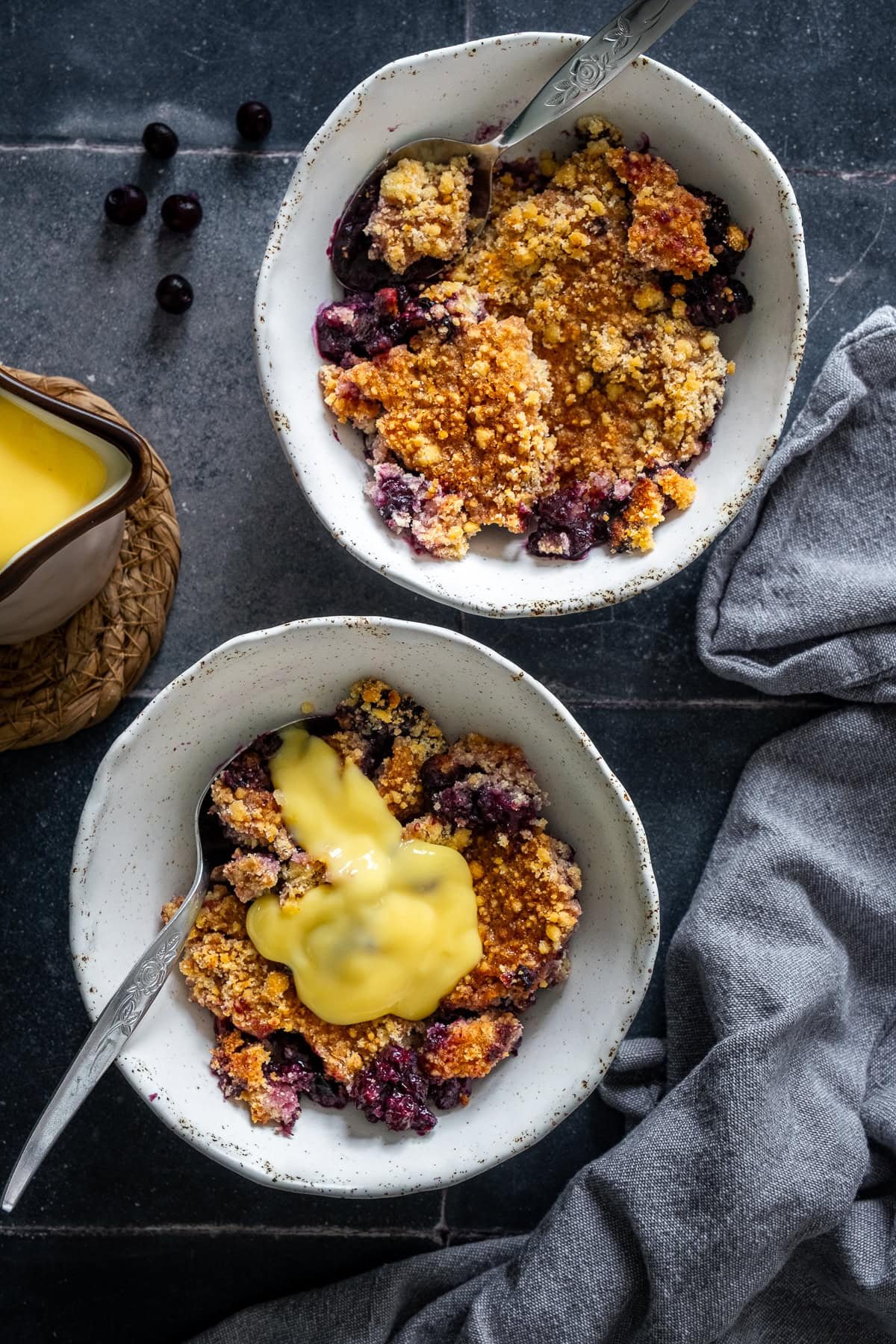 Overhead shot of two bowls of blueberry crumble. One had custard on top.