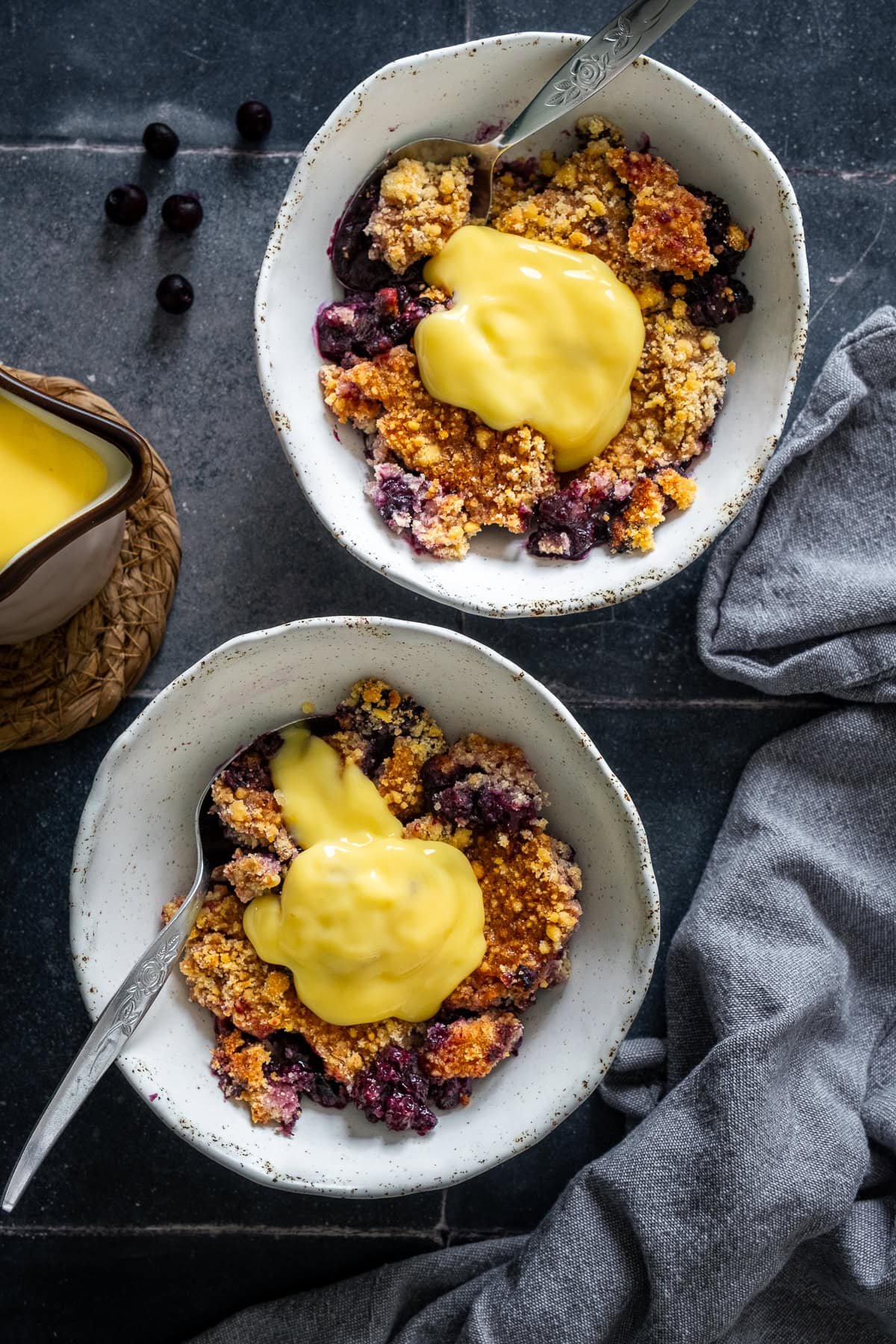 Overhead shot of of 2 bowls of Easy Blueberry Crumble with custard on top.