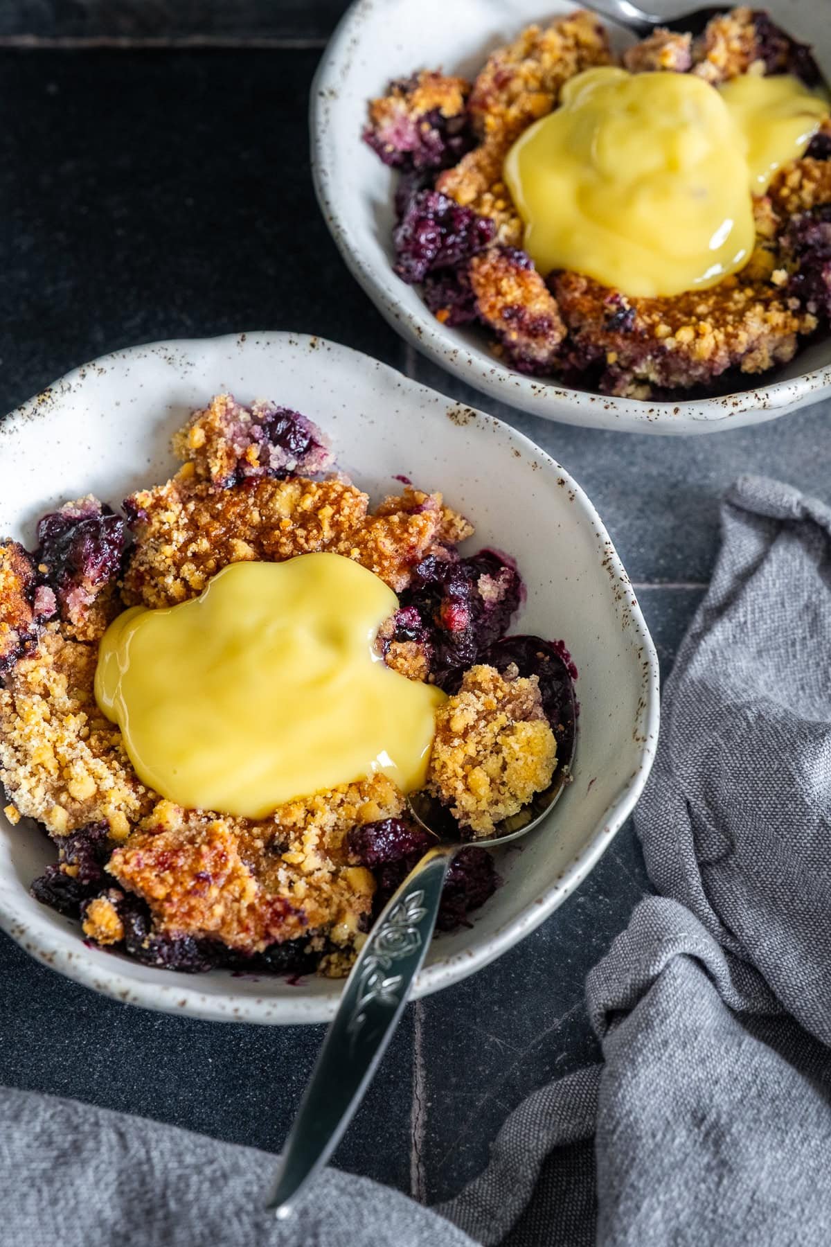 Closeup of 2 bowls of Easy Blueberry Crumble with custard on top.