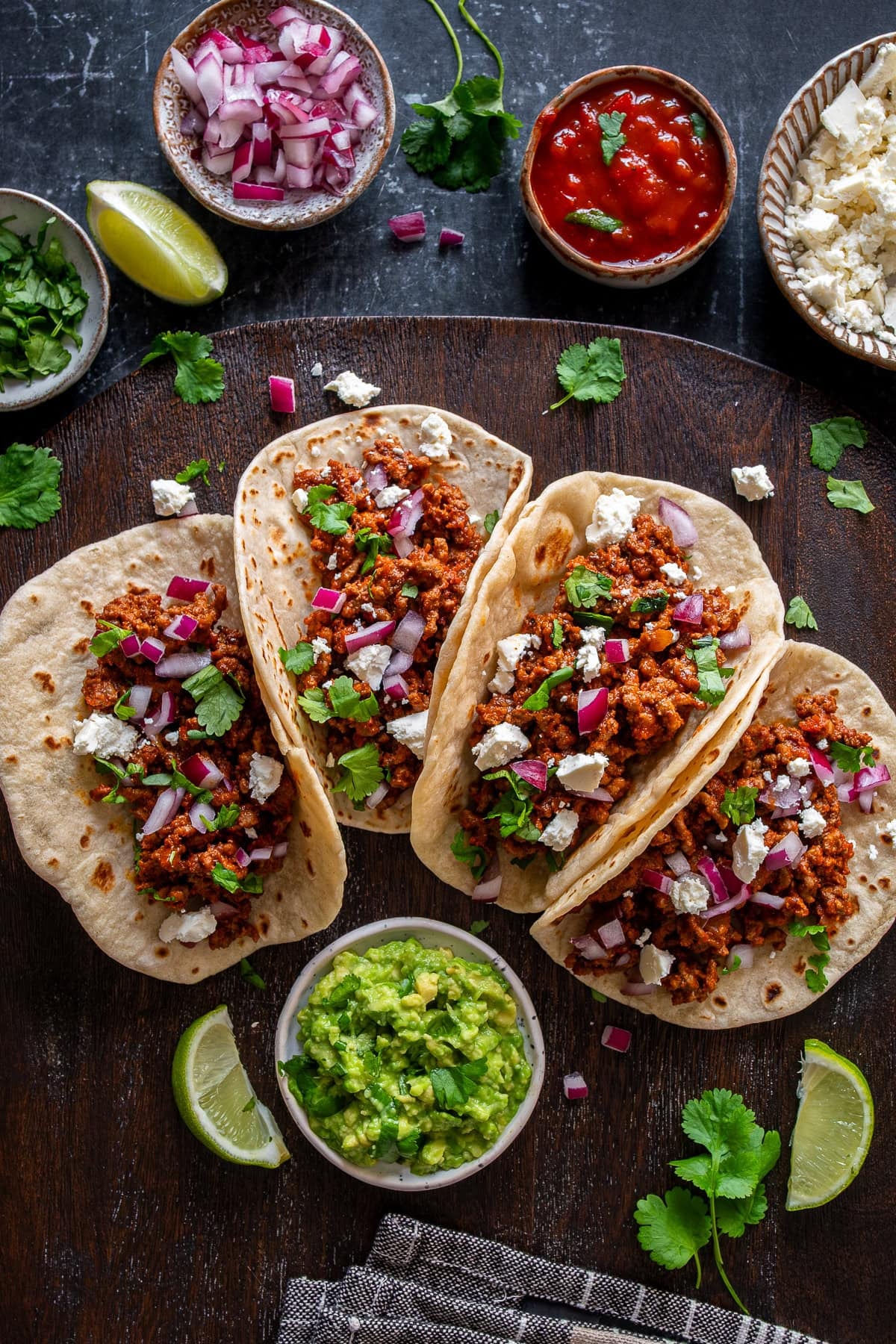 Overhead shot of 4 Easy Beef Mince Tacos with feta cheese, diced red onions, fresh coriander, lime quarters, salsa and guacamole.