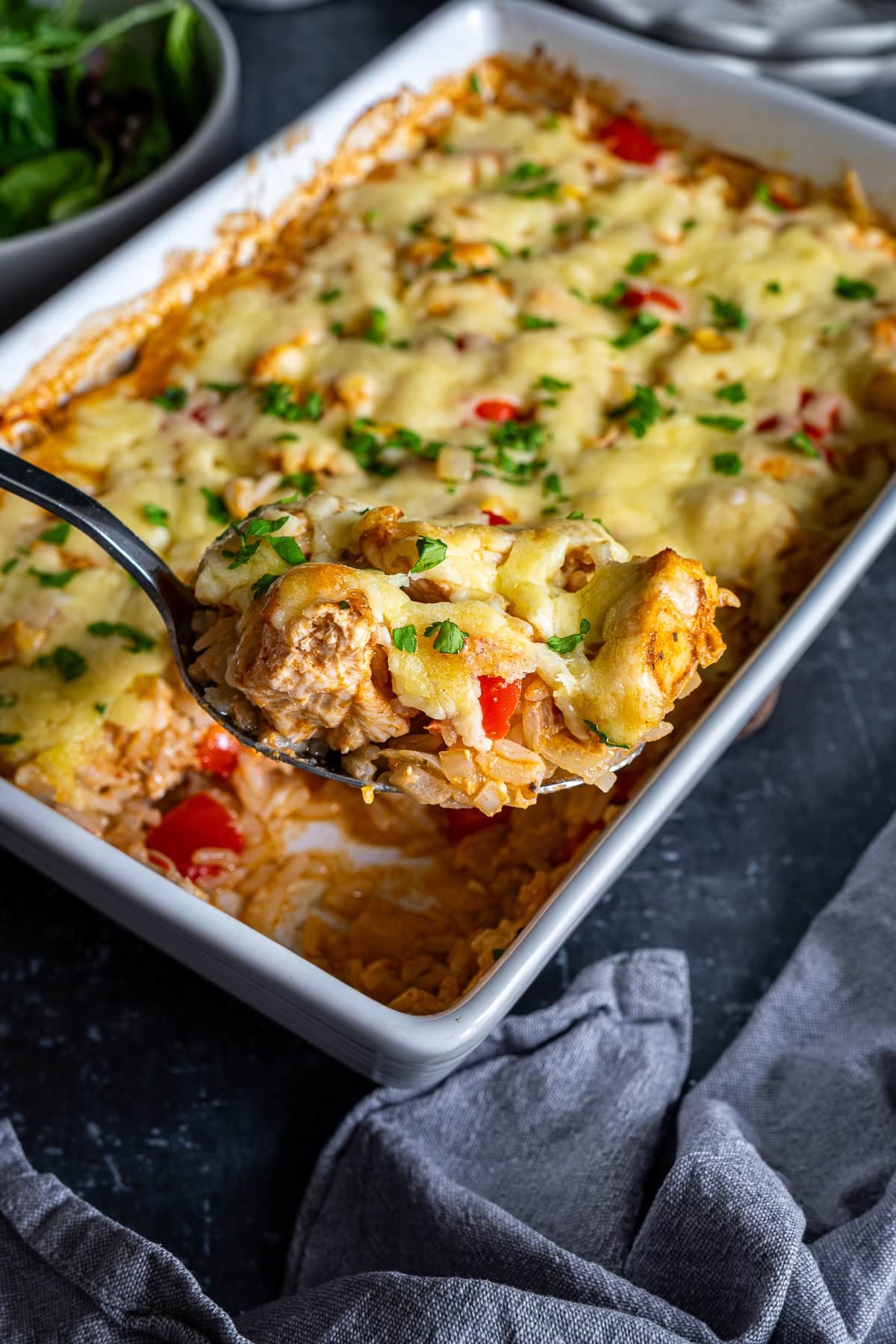 Closeup of a serving spoon with Chicken Fajita Rice Bake on it. The full oven dish of Chicken Fajita Rice Bake is in the background.