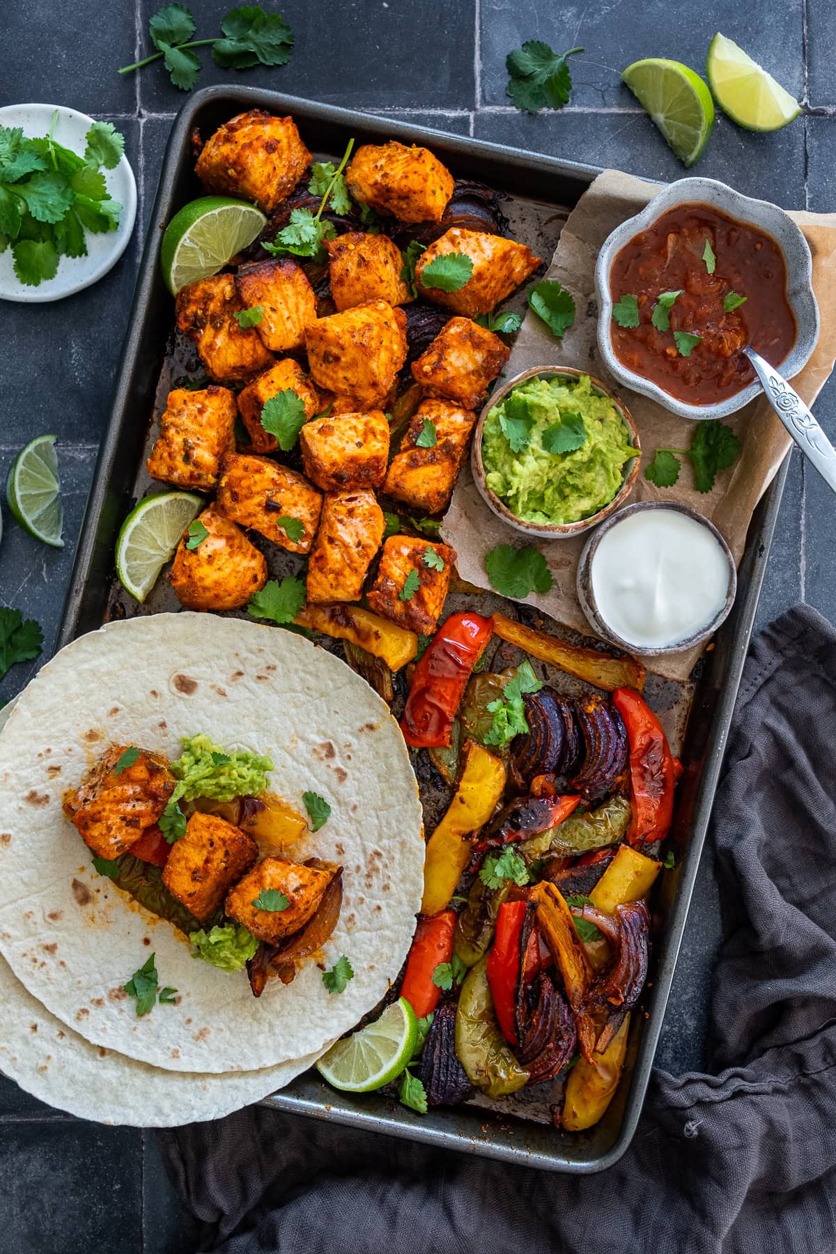 Overhead shot of One Pan Salmon Fajitas in the oven tray, served with salsa, guacamole, sour cream, tortillas, coriander and limes.