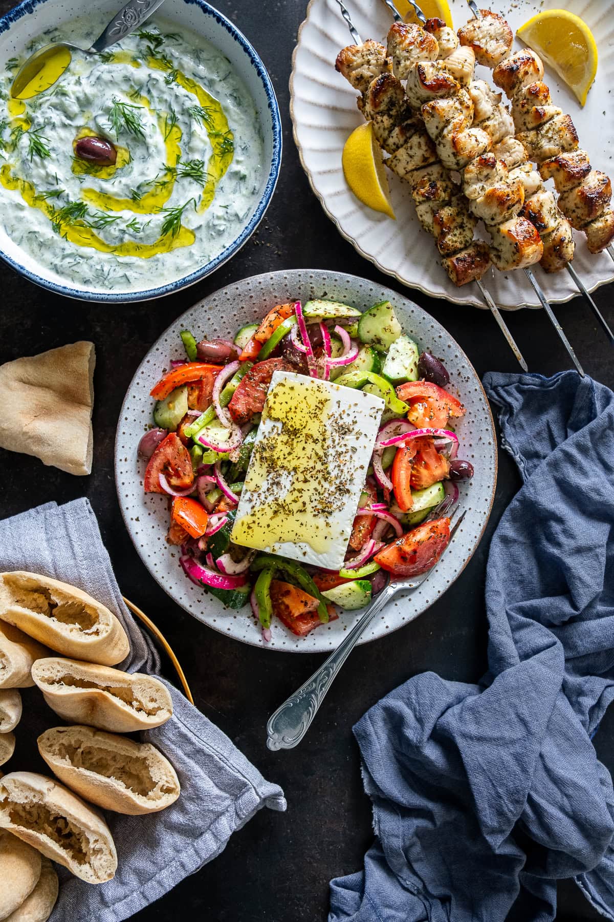 Overhead shot of Authentic Greek Salad surrounded by tzatziki, pittas and chicken souvlaki
